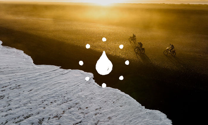 A dramatic aerial landscape photo at sunset showing a beach with the tide coming in, and three cyclists riding in the distance. A white water-drop icon with surrounding dots is overlaid near the center.